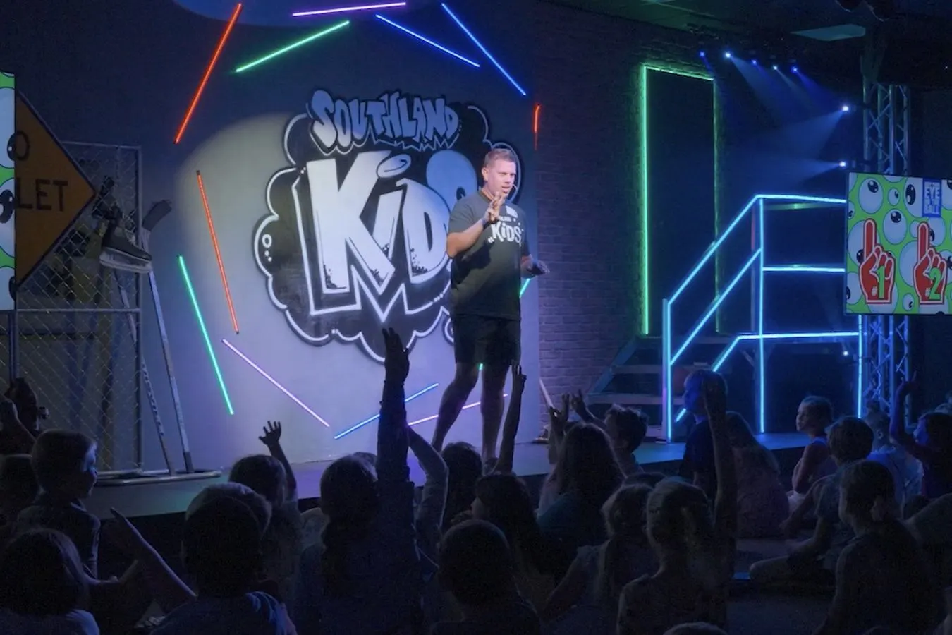 A Southland Kids leader teaching on a colorful stage while kids raise their hands